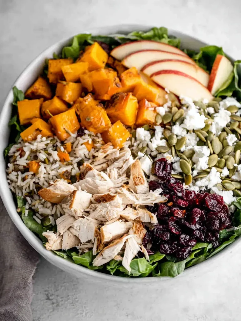 Overhead view of a white bowl filled with a colorful salad. Ingredients include roasted butternut squash, shredded chicken, brown rice, dried cranberries, chopped apples, pumpkin seeds, and feta cheese over green leaves.