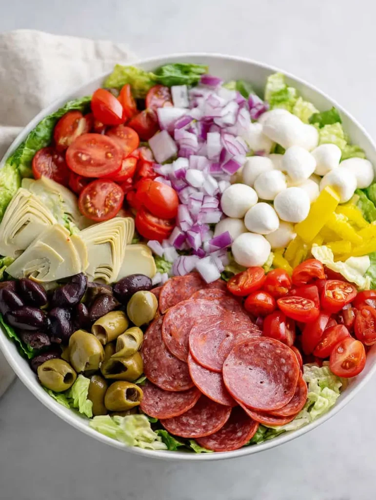 An overhead view of a large, colorful Italian chopped salad in a white bowl, featuring lettuce, pepperoni, salami, mozzarella, tomatoes, olives, red onion, artichokes, and pepperoncini.