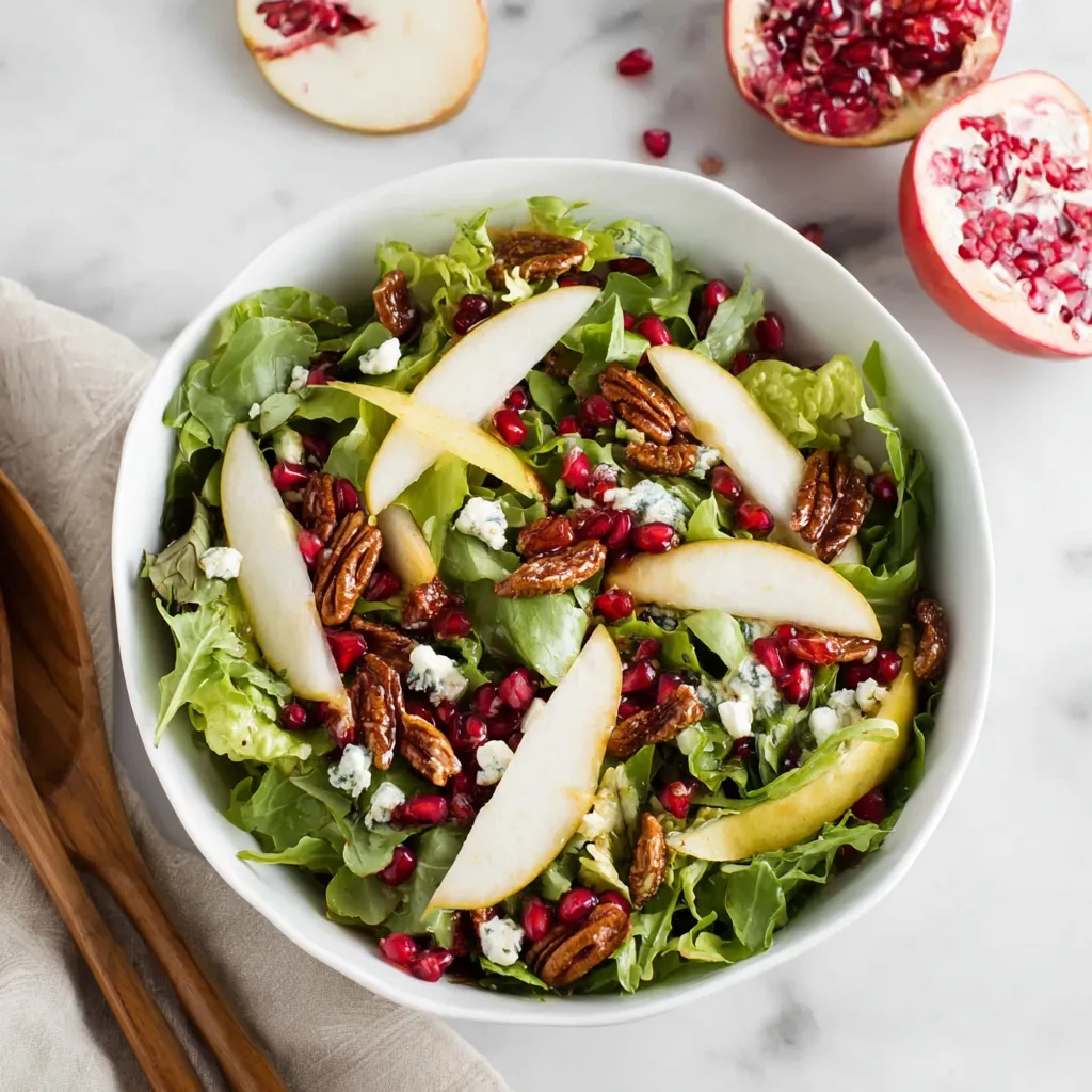 Overhead view of a salad with mixed greens, sliced pears, pecans, pomegranate, and crumbled cheese in a white bowl. A pear half, pomegranate half, and tongs are on a white marble surface nearby.
