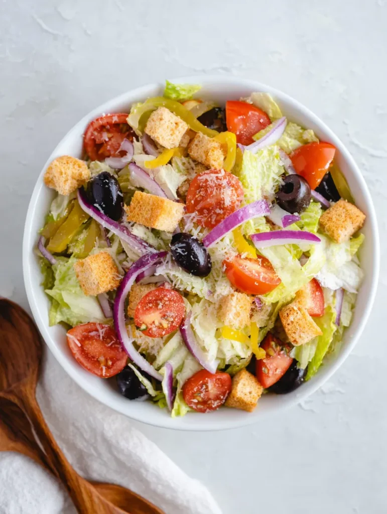 Overhead view of a colorful garden salad in a white bowl. It contains shredded lettuce, sliced tomatoes, black olives, red onion, yellow pepperoncini, croutons, and grated cheese. A white napkin and wooden utensils are partially visible on the left.
