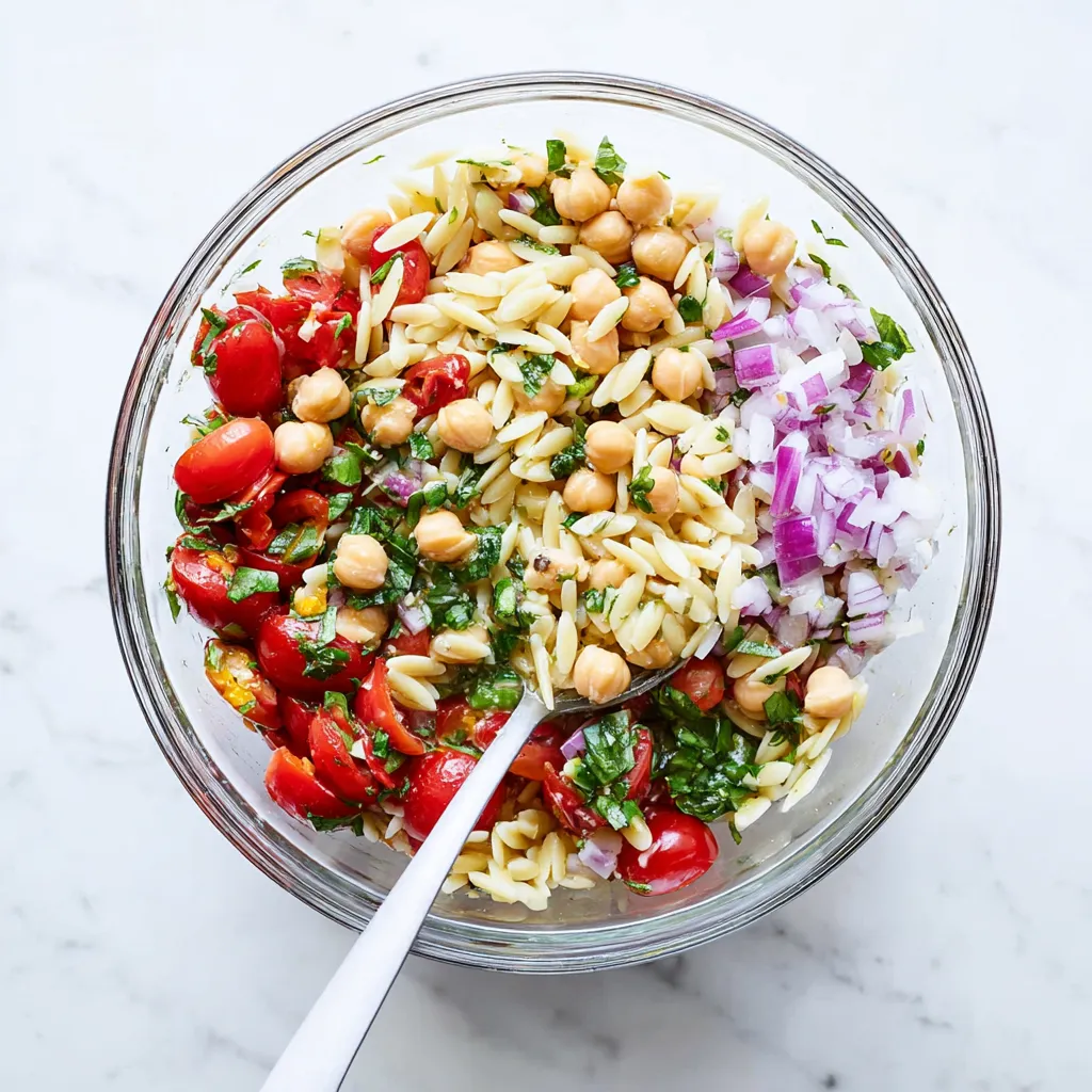 Overhead view of an orzo pasta salad in a clear glass bowl, featuring orzo pasta, chickpeas, halved red cherry tomatoes, diced red onion, and green herbs. A white spoon rests in the salad on a white marble surface.