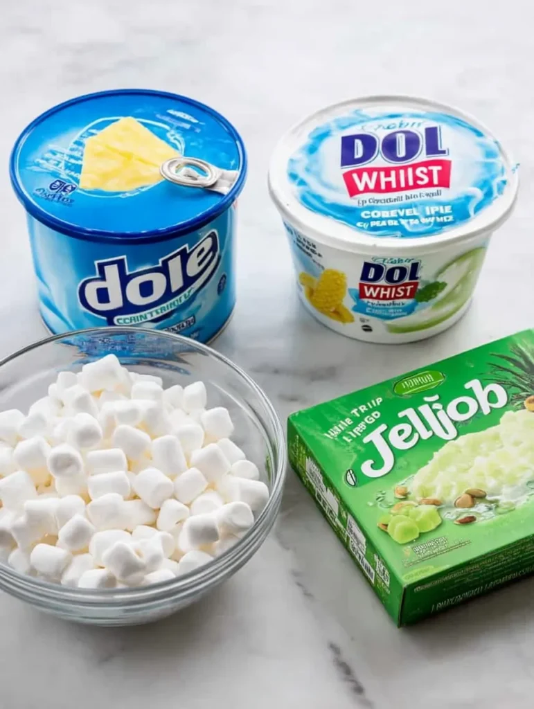 Overhead shot of Watergate Salad ingredients: a can of Dole crushed pineapple, Cool Whip tub, Jell-O pistachio pudding mix, and a bowl of marshmallows on white marble.