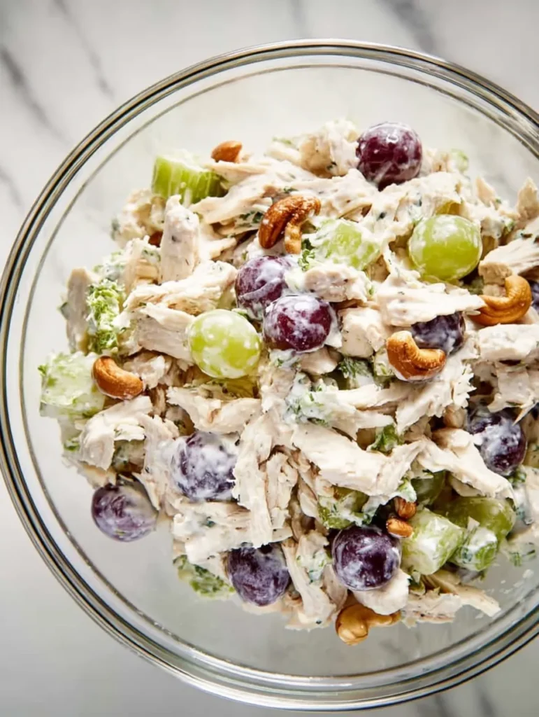 Overhead view of a chicken salad in a clear glass bowl, containing shredded chicken, halved grapes, diced celery, and cashews with a creamy dressing, on a white marble countertop.