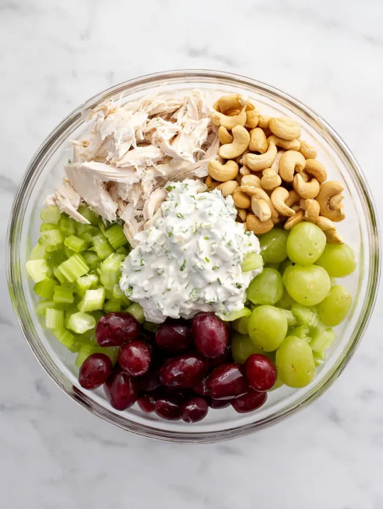 Overhead view of chicken salad ingredients in a clear bowl. Includes shredded chicken, cashews, halved red grapes, diced celery, and creamy dressing.