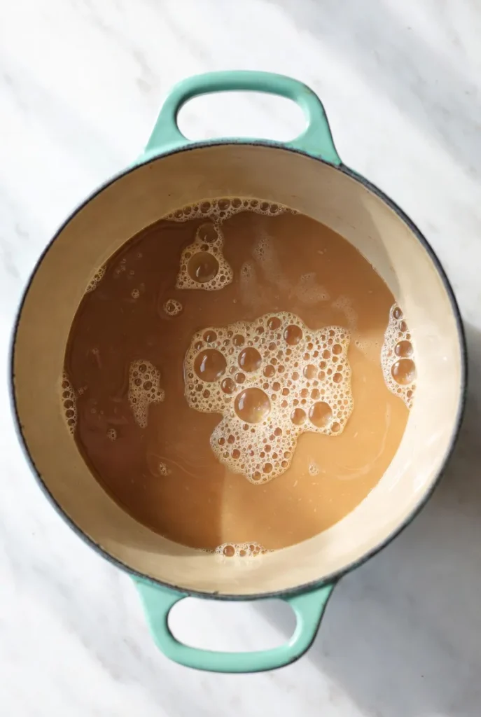 Overhead view of a light blue enameled cast iron pot with a creamy interior, holding a simmering, foamy light brown liquid on white marble.